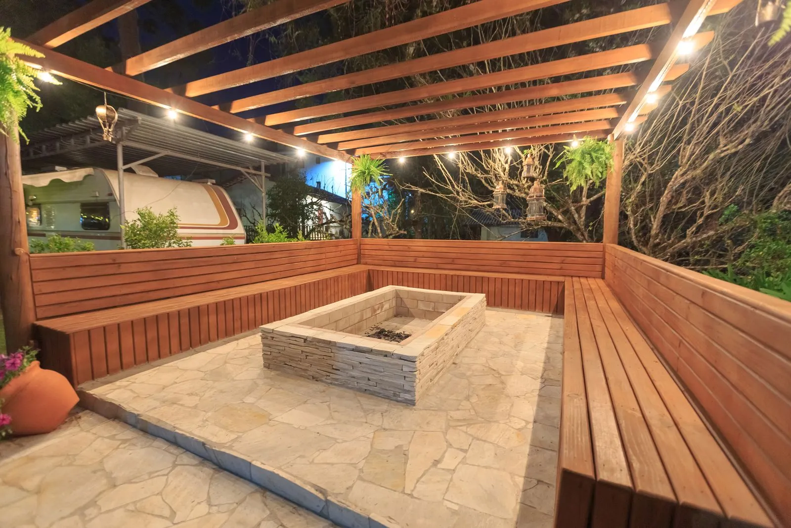 A wood pergola with built-in benches around a stone fire pit at night